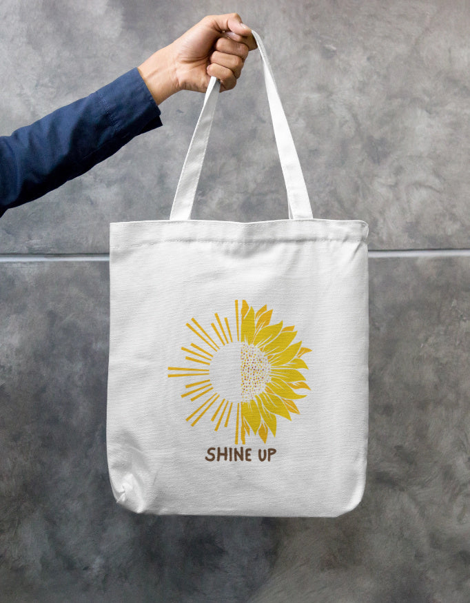 Tote bag with a sunflower design and 'Shine Up' text held by a person against a gray background